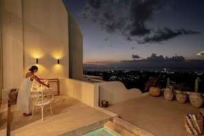 A terrace with a table and chairs, a view of the city at dusk, and potted plants. - Villa Mabu (Uluwatu)