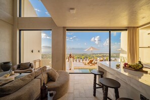 A modern living room with a large window, a sofa, a coffee table, and a bar area with a fruit bowl. - Villa Mabu (Uluwatu)
