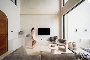 A modern living room with a large window, a flat-screen TV, and a sofa. - Villa Mabu (Uluwatu)