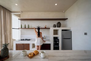 A modern kitchen with marble countertops, a built-in oven, and a refrigerator. There are wine bottles and glasses on the shelves, and a pineapple on the counter. - Villa Mabu (Uluwatu)