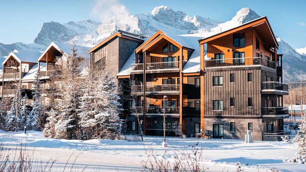 A multi-story hotel with a wooden facade, balconies, and a snowy mountain backdrop. - Hidden Cirque Hydeaway at Ascent (Canmore)