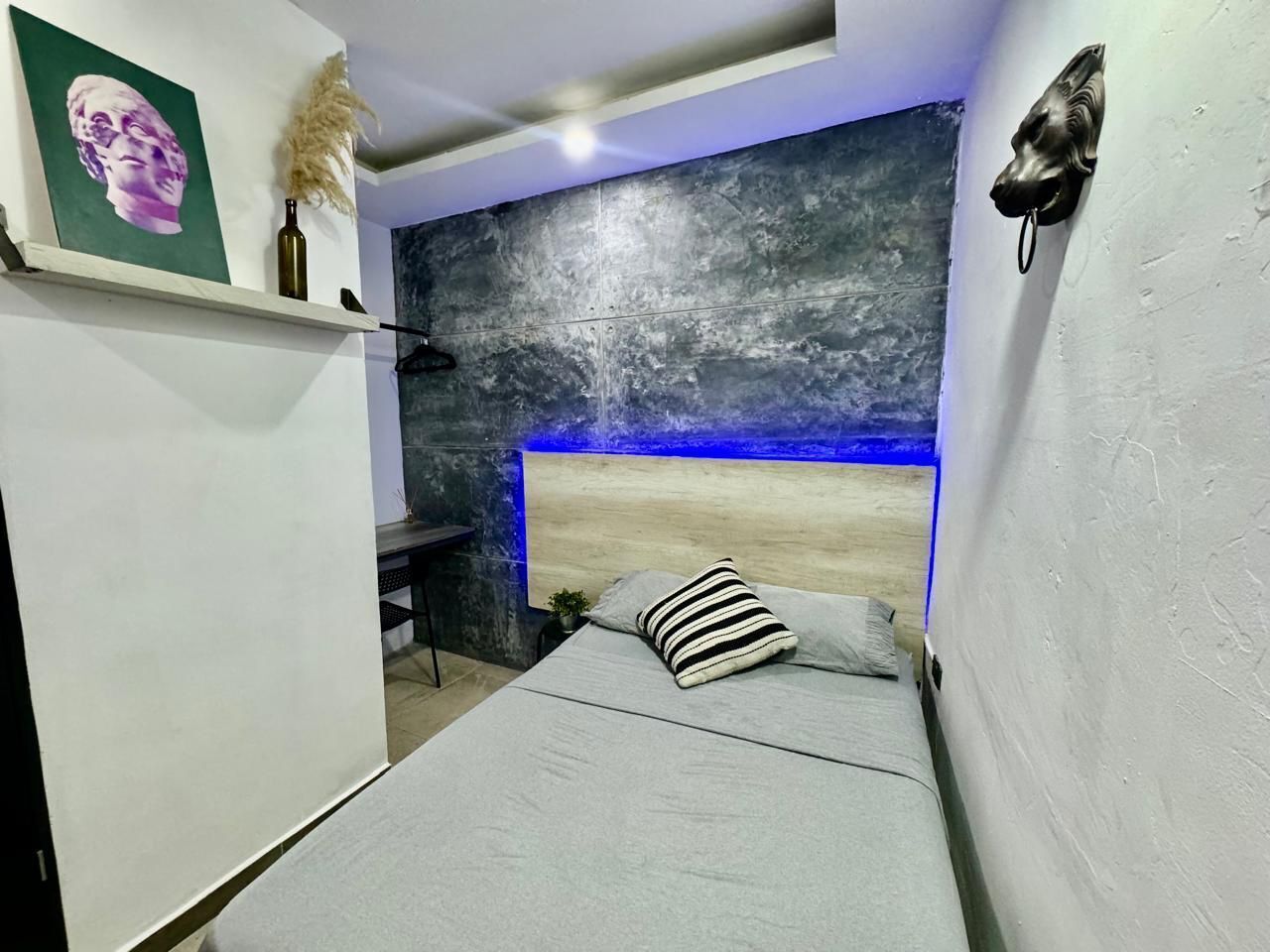 Un dormitorio con cama, cabecera con iluminación azul, escultura en la pared y estante con una botella y un objeto decorativo.