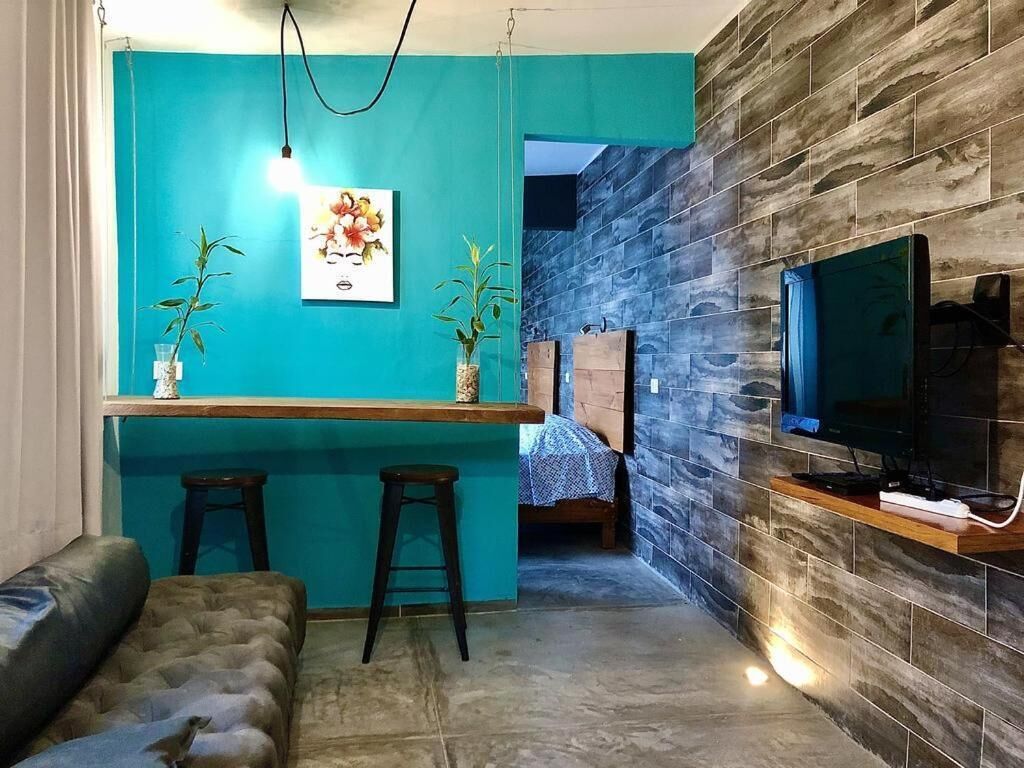 A compact living space with a teal accent wall, a small bar area, a sofa, a TV mounted on a stone wall, and a bed in the background.