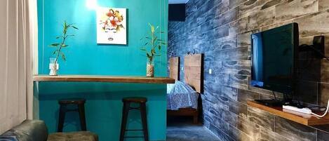 A compact living space with a teal accent wall, a small bar area, a sofa, a TV mounted on a stone wall, and a bed in the background.