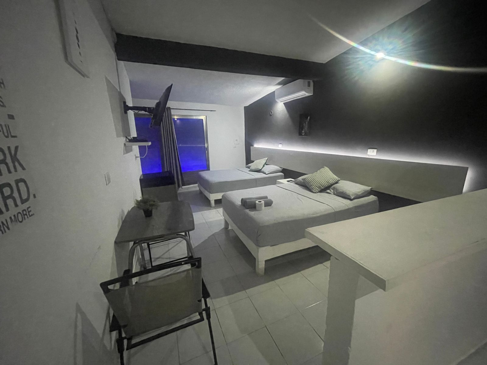 A modern, minimalist bedroom with a large bed, a small desk, and a chair. There is a blue-lit room in the background.