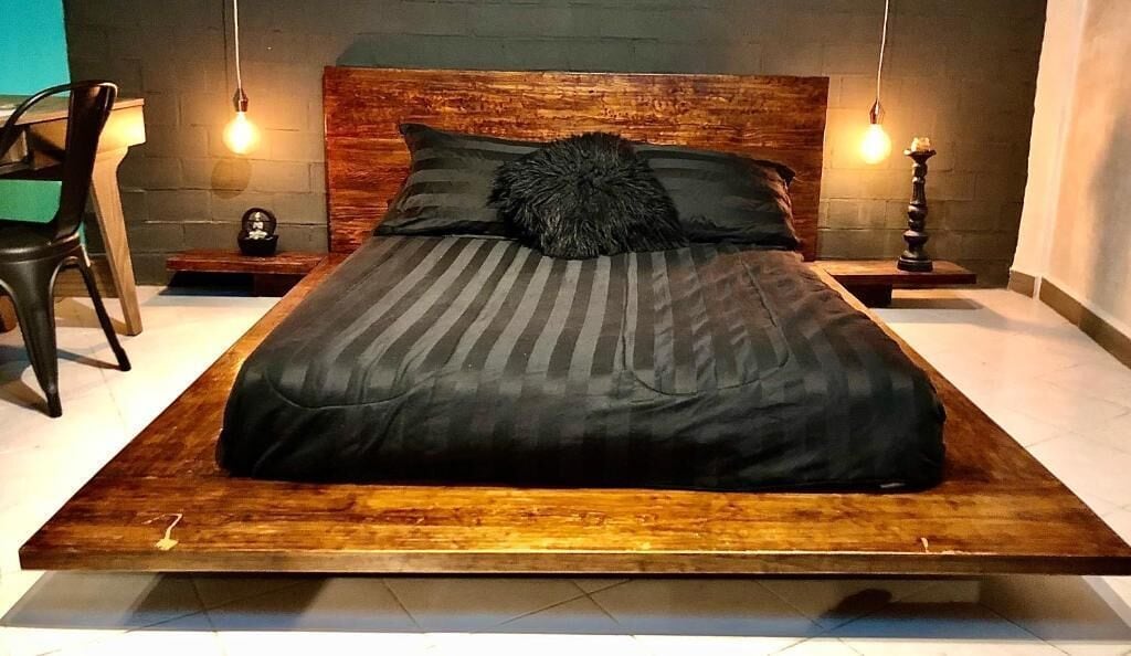Una cama moderna con estructura de madera y colcha a rayas oscuras. Hay dos mesitas de noche con objetos decorativos y una silla a la izquierda.