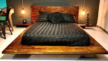 Una cama moderna con estructura de madera y colcha a rayas oscuras. Hay dos mesitas de noche con objetos decorativos y una silla a la izquierda.