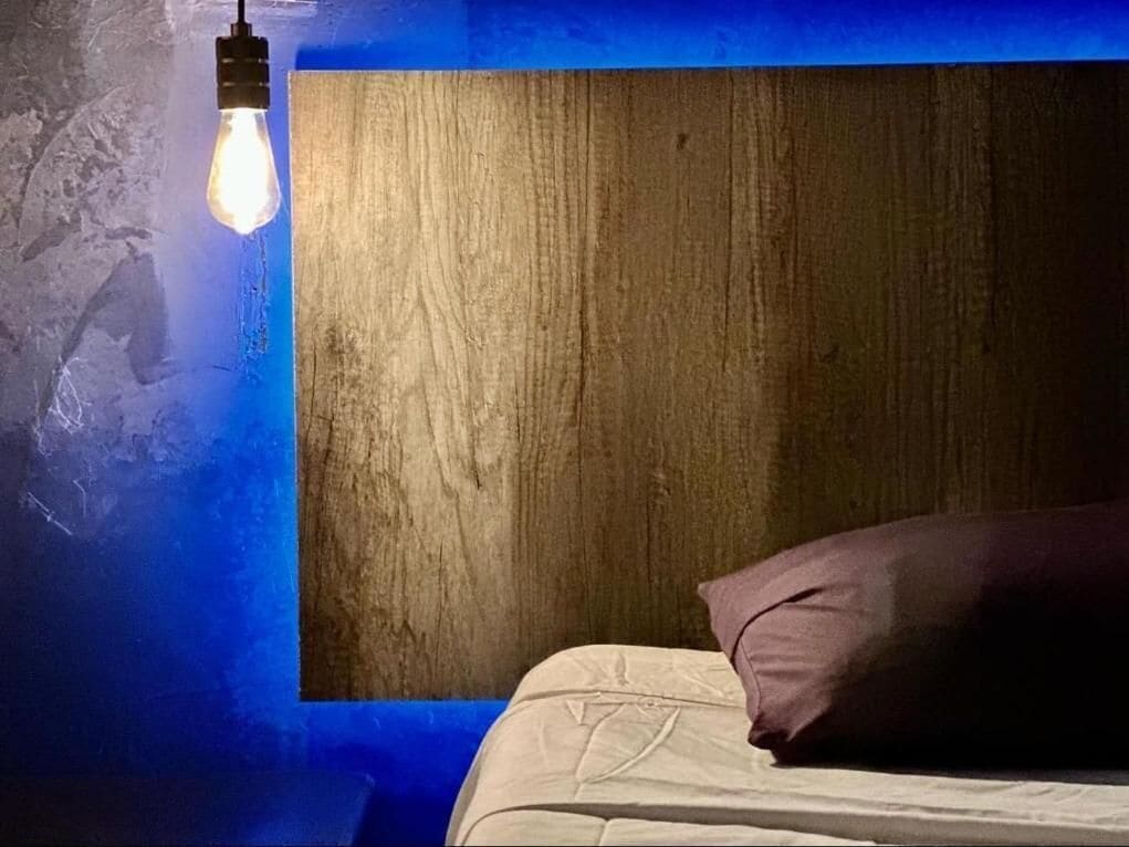 A bedroom with a wooden headboard, a single bed with a white mattress and a purple pillow, and a light bulb hanging from the ceiling.