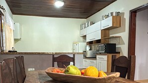 A wooden dining table with a fruit bowl in the center, a kitchen area with cabinets and appliances in the background, and a wooden ceiling. - Country house with pool and beautiful gourmet area near Holambra. (Artur Nogueira)