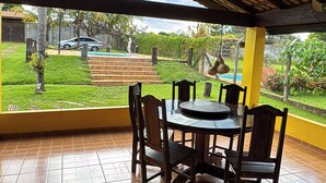 An outdoor patio with a round table and chairs, a thatched roof, and a view of a swimming pool and garden. - Country house with pool and beautiful gourmet area near Holambra. (Artur Nogueira)