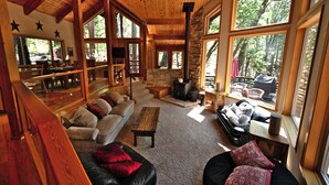 A cozy cabin interior with a stone fireplace, wooden furniture, and large windows offering a view of the forest.