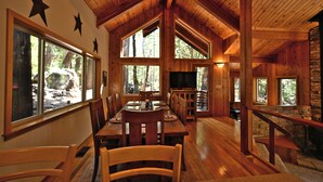 A spacious dining area with wooden tables and chairs, large windows offering views of the surrounding forest, and a stone fireplace.