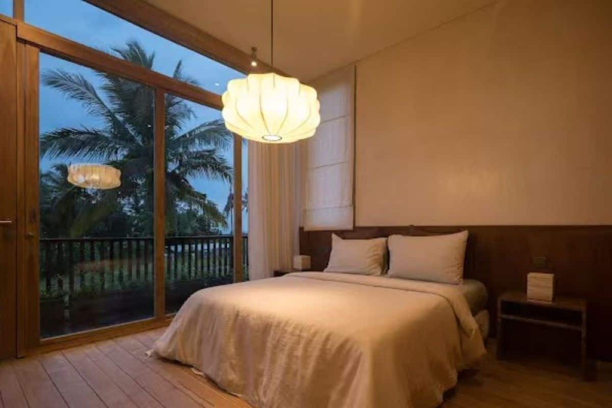 A bedroom with a large bed, a nightstand, a hanging light fixture, and a view of palm trees through a sliding glass door.