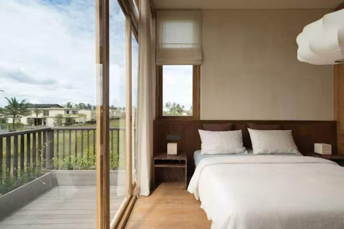 A bedroom with a large window, wooden flooring, a bed with white bedding, and a nightstand with a lamp.