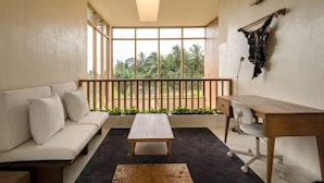A room with a sofa, a table, and a desk. There is a window with a view of greenery outside. - KOO villa (Pejeng Kawan)