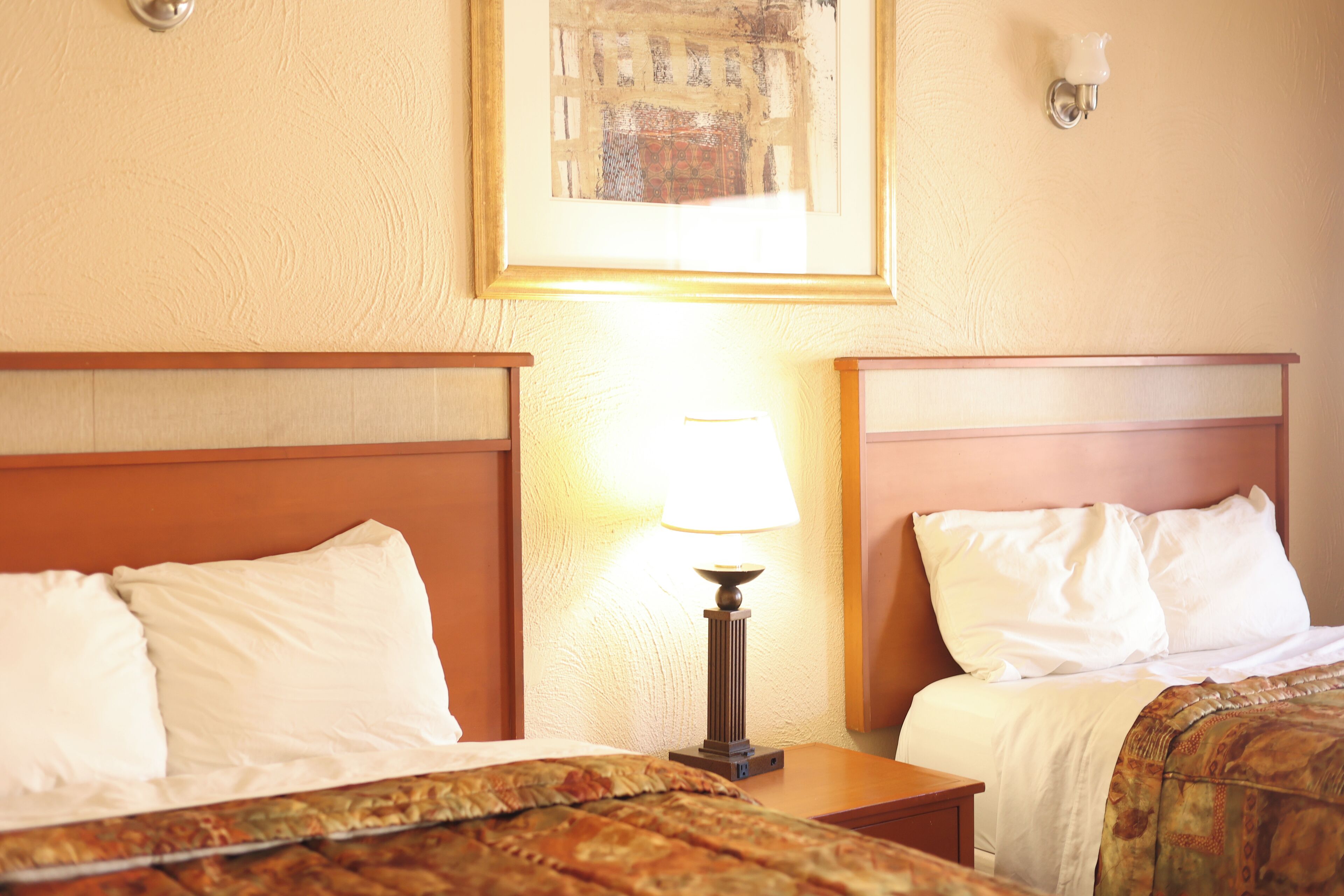 Two beds with wooden headboards, a nightstand with a lamp, and a framed picture on the wall.