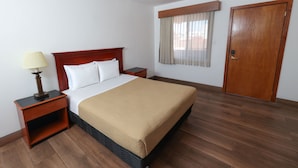 Een hotelkamer met een houten bed, een nachtkastje met lamp, een houten deur en een raam met gordijnen.