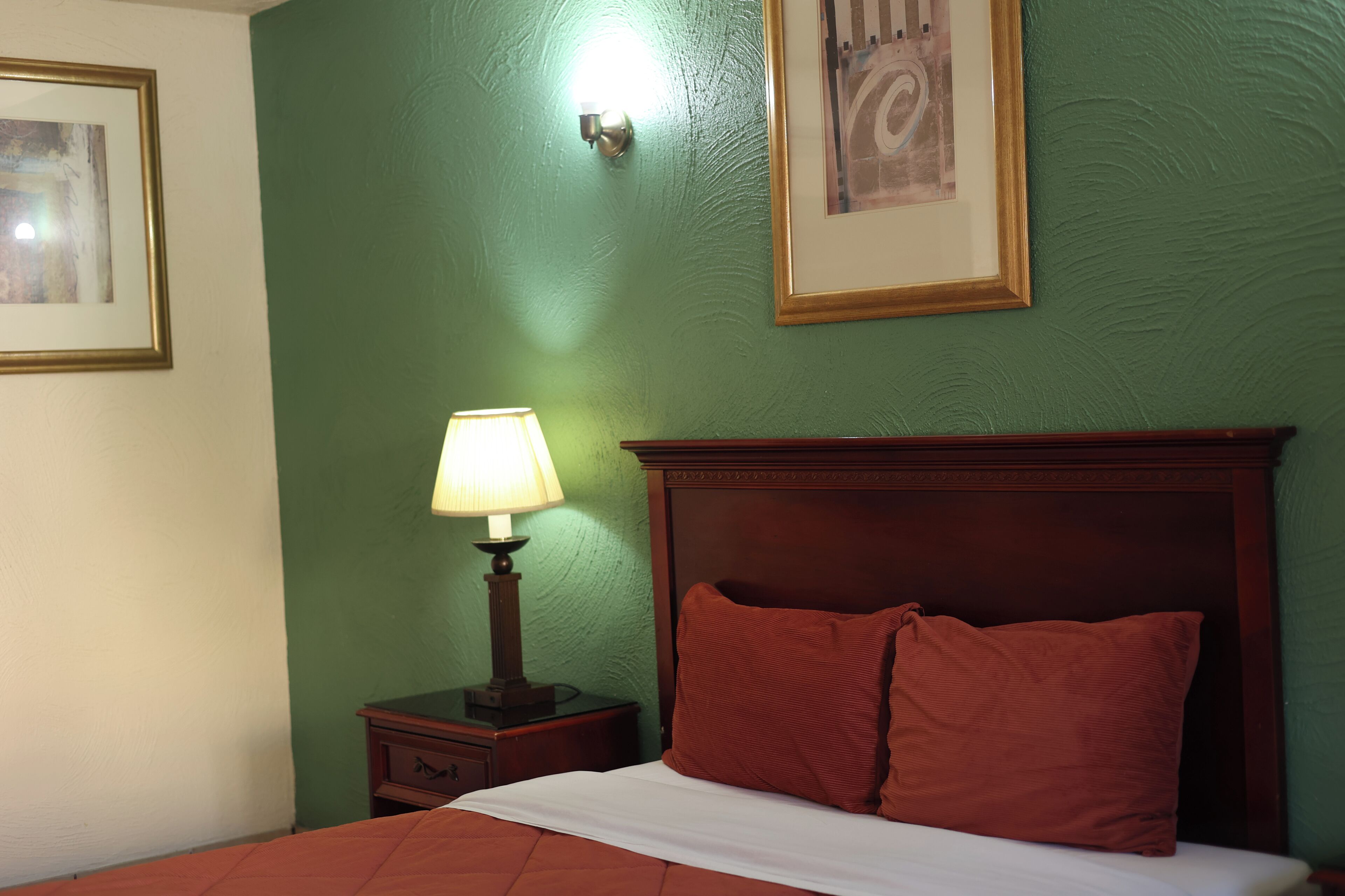Una cama bien hecha con un cojín rojo, una lámpara de noche y un cuadro en la pared.