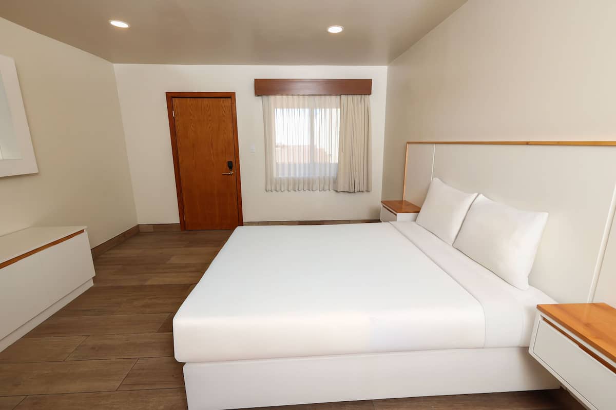 Ein Hotelzimmer mit einem großen Bett, einem Holzkopfstück, einem Nachttisch, einem Beistelltisch, einer Holzbank, einer Holztür und einem Fenster mit Vorhängen.