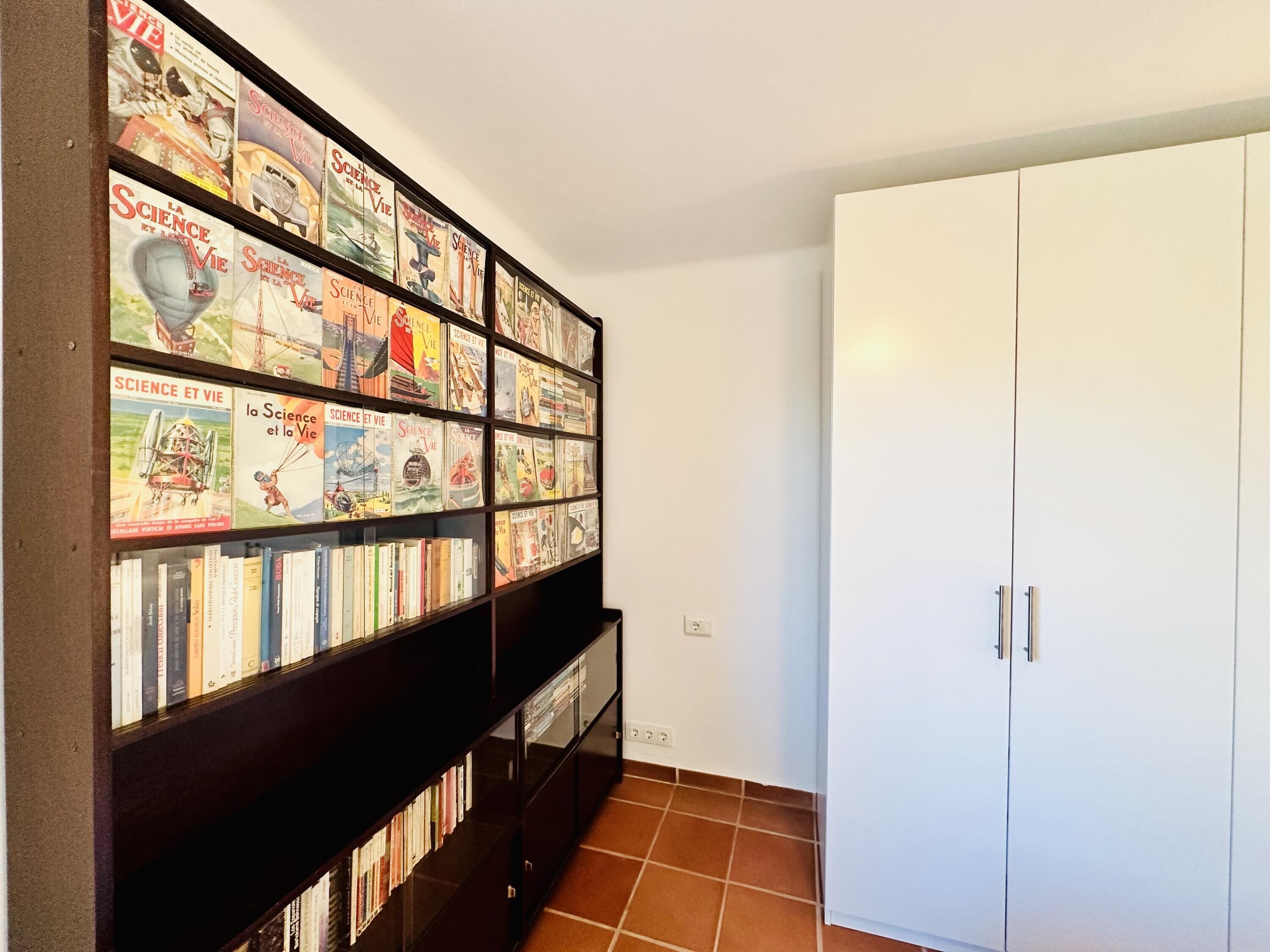 A room with a bookshelf filled with books and magazines, a white wardrobe, and a tiled floor.