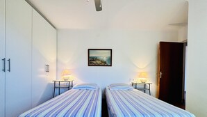A room with two single beds, each with a striped bedspread, two bedside tables with lamps, a painting on the wall, and a door to the right. - BARCA.Apartment with direct Beach Access (ROSES)
