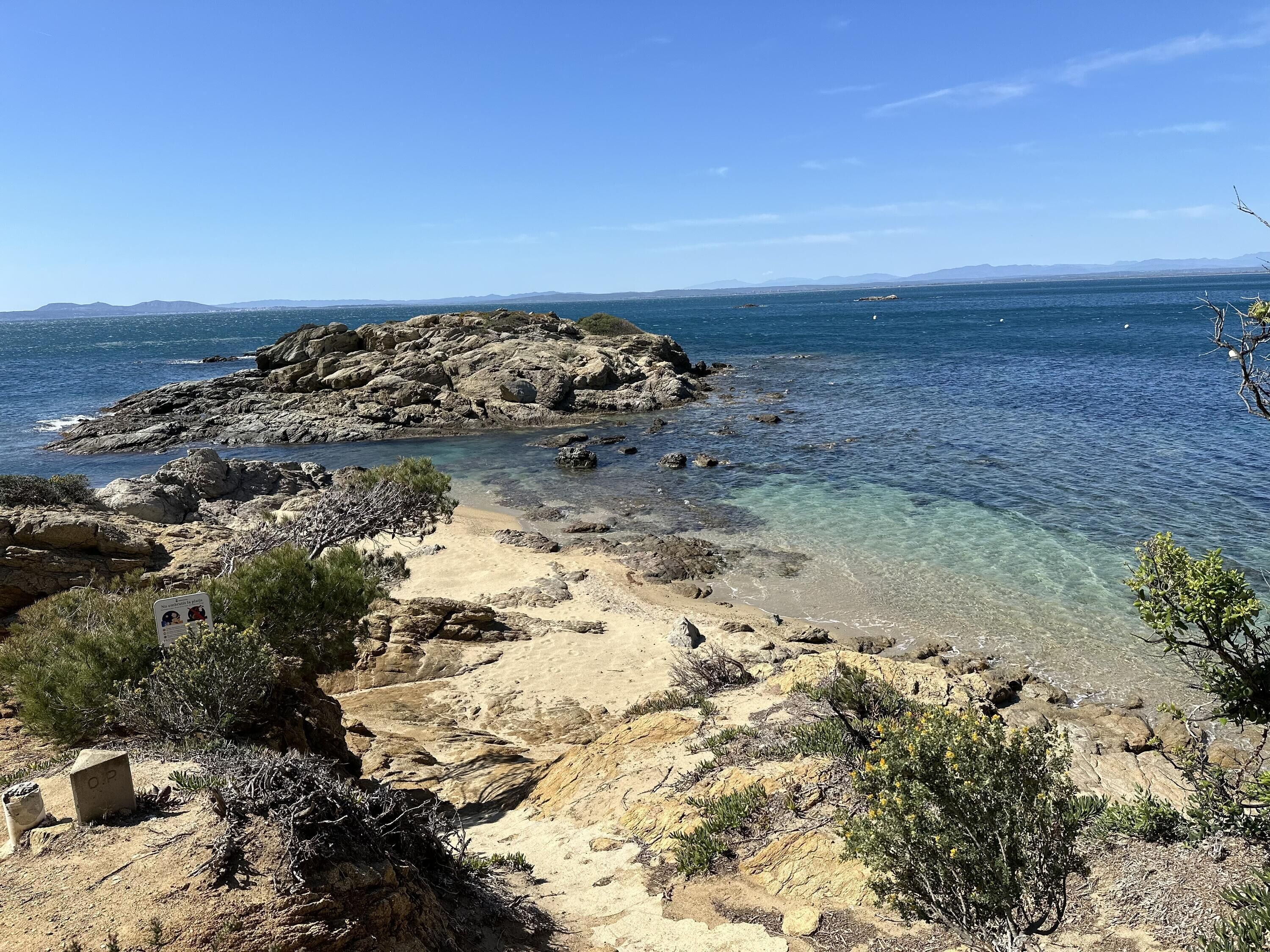 A rocky beach with clear water, a sandy path, and sparse vegetation.