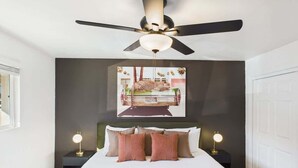 Ein Schlafzimmer mit einem Bett, Deckenventilator, Wandbild, Nachttischlampen und einer Tür.