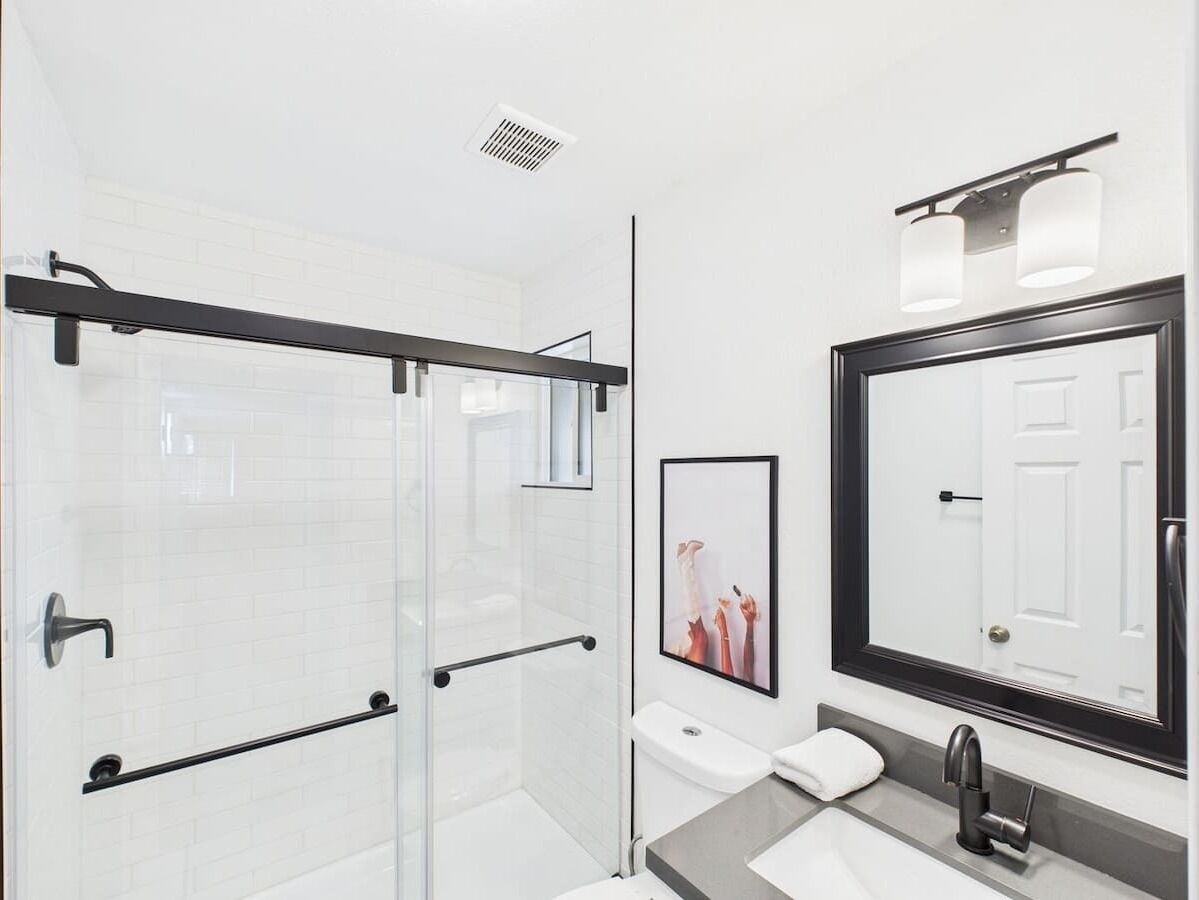 A modern bathroom with a glass shower enclosure, a white toilet, a sink with a mirror, and a framed picture on the wall.
