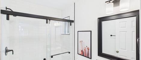 A modern bathroom with a glass shower enclosure, a white toilet, a sink with a mirror, and a framed picture on the wall.