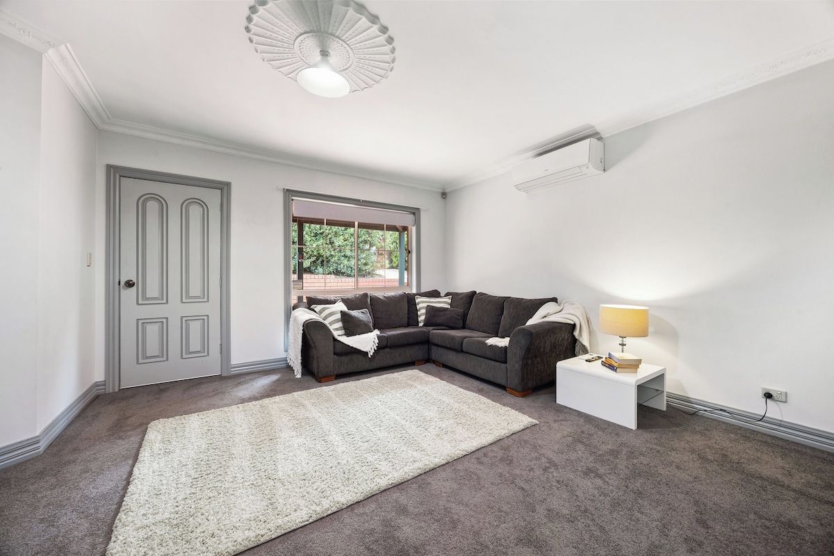 A living room with a sectional sofa, a white rug, a side table with a lamp, and a window with a view of greenery.