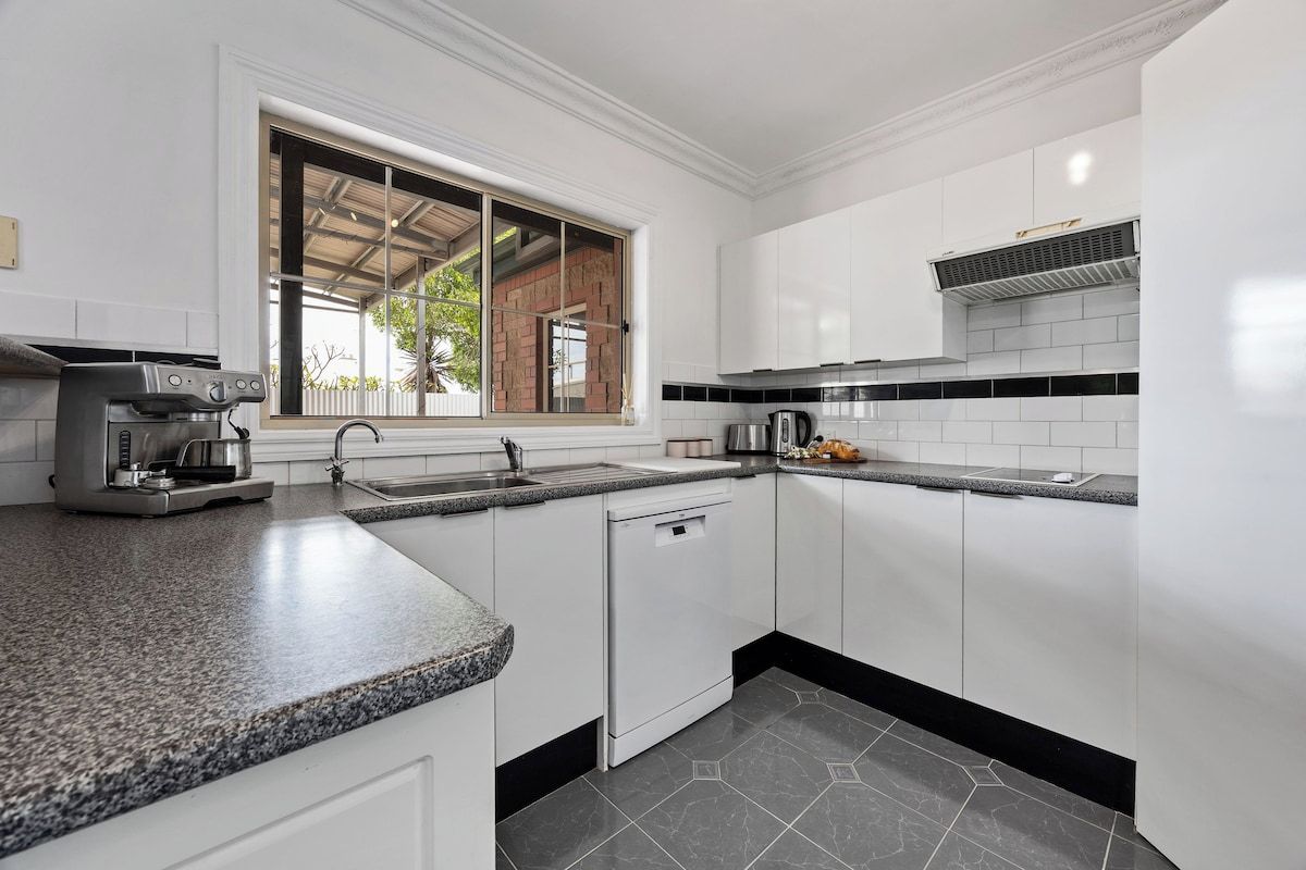 A kitchen with white cabinets, a granite countertop, a coffee maker, and a window with a view of a brick building.