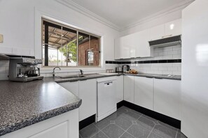 A kitchen with white cabinets, a granite countertop, a coffee maker, and a window with a view of a brick building. - The Laneway | 3BR - Families & Crews · Walk To CBD (Port Lincoln)