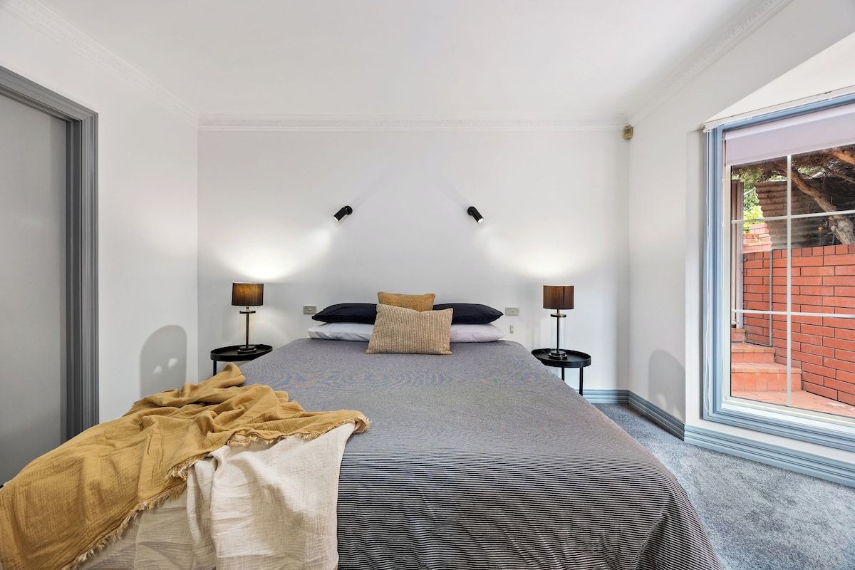 Un dormitorio con una cama grande, dos mesitas de noche con lámparas, una ventana con vista a un muro de ladrillo y una puerta a la izquierda.