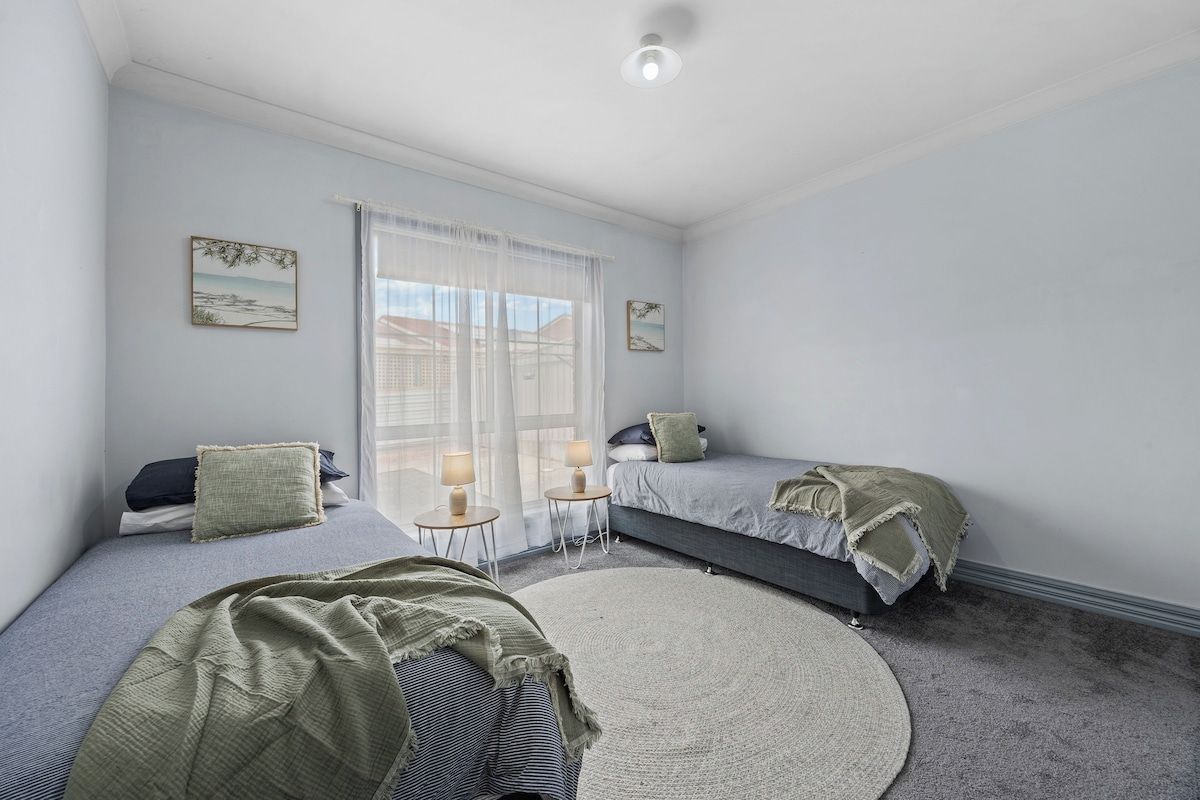 A bedroom with two single beds, a large window with sheer curtains, and a round rug on the floor.