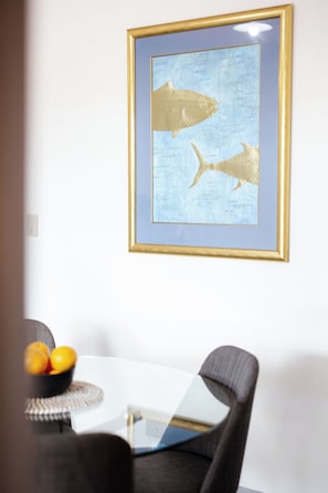A framed artwork of a fish on a wall, a white table with a bowl of fruit, and a chair.