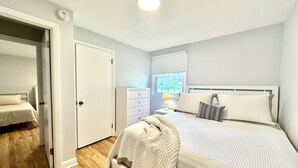 Un dormitorio con dos camas, un tocador, una mesita de noche con lámpara, una ventana con persianas y una puerta que da a otra habitación.