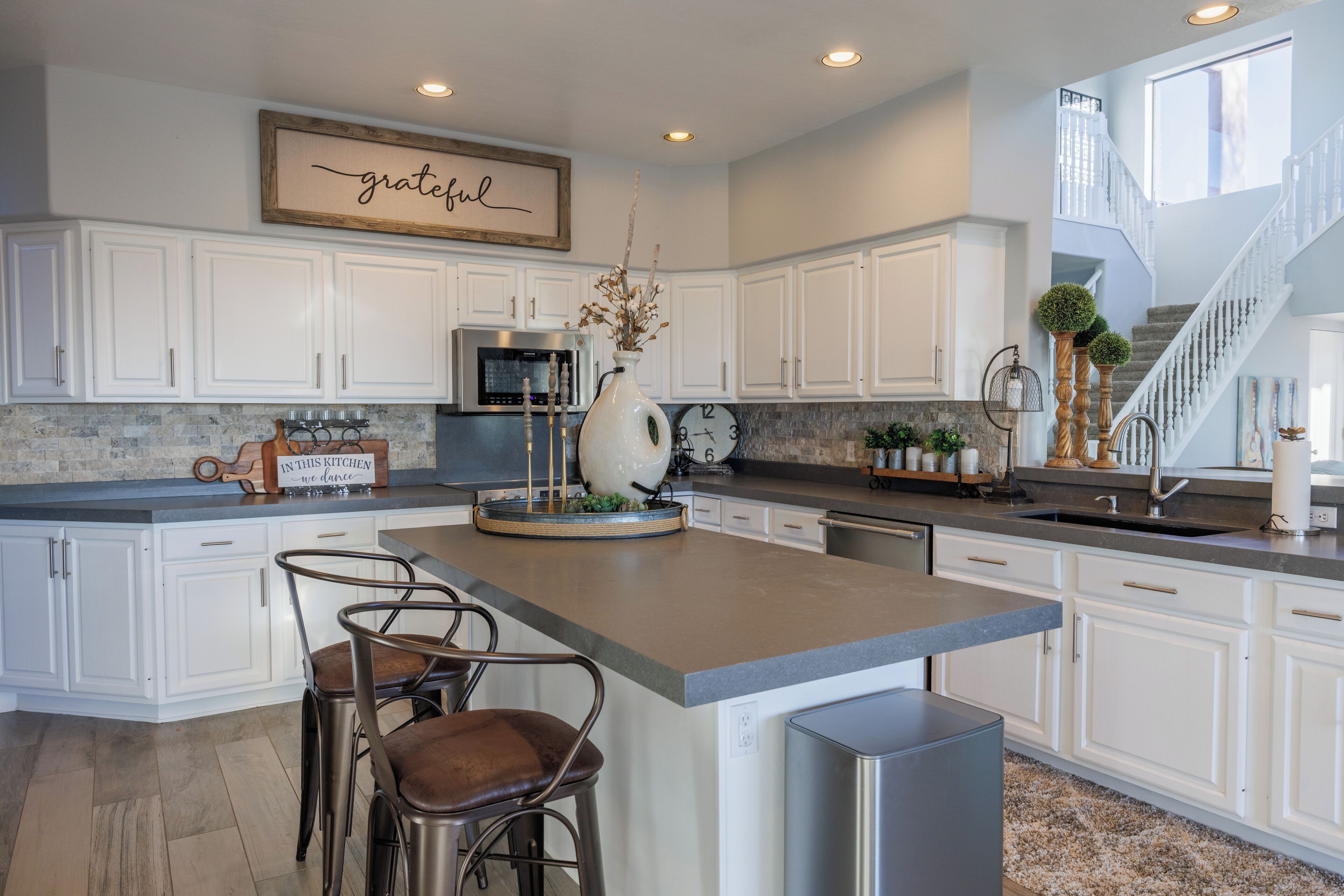 A spacious kitchen with white cabinets, a central island with bar stools, a clock, and a decorative sign above the cabinets.