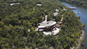 Une vue aérienne d’une grande maison à plusieurs étages, entourée d’une forêt dense, avec une jetée qui s’avance dans un lac.
