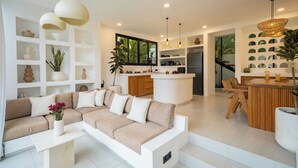 A modern living room with a beige sectional sofa, white coffee table, and a kitchen area with a central island. - Villa Joann (Uluwatu)
