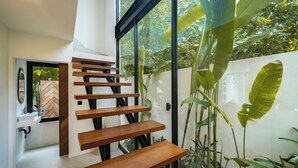 A modern staircase with wooden steps and black railings, leading to a room with a white wall and a large window overlooking greenery. - Villa Joann (Uluwatu)