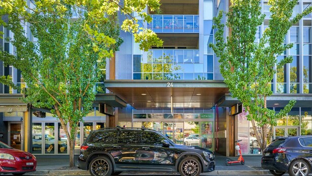 A modern building with a glass facade, numbered 24, featuring a covered entrance and a parking area with cars. - Spacious Studio in a Prime Braddon Location (Braddon)