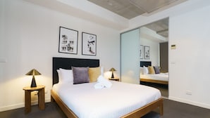 A modern hotel room with a large bed, two bedside tables with lamps, a large mirror, and framed pictures on the wall. - Spacious Studio in a Prime Braddon Location (Braddon)