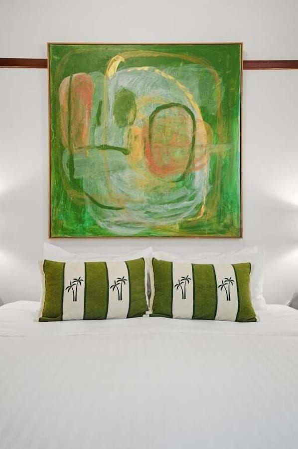 A bedroom with a large abstract painting on the wall, a bed with white bedding, and two pillows with palm tree designs.