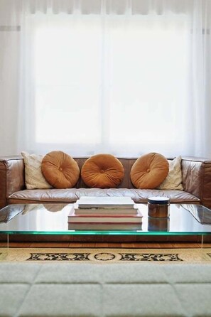 A living room with a leather sofa, three round cushions, and a glass coffee table with books and a candle.