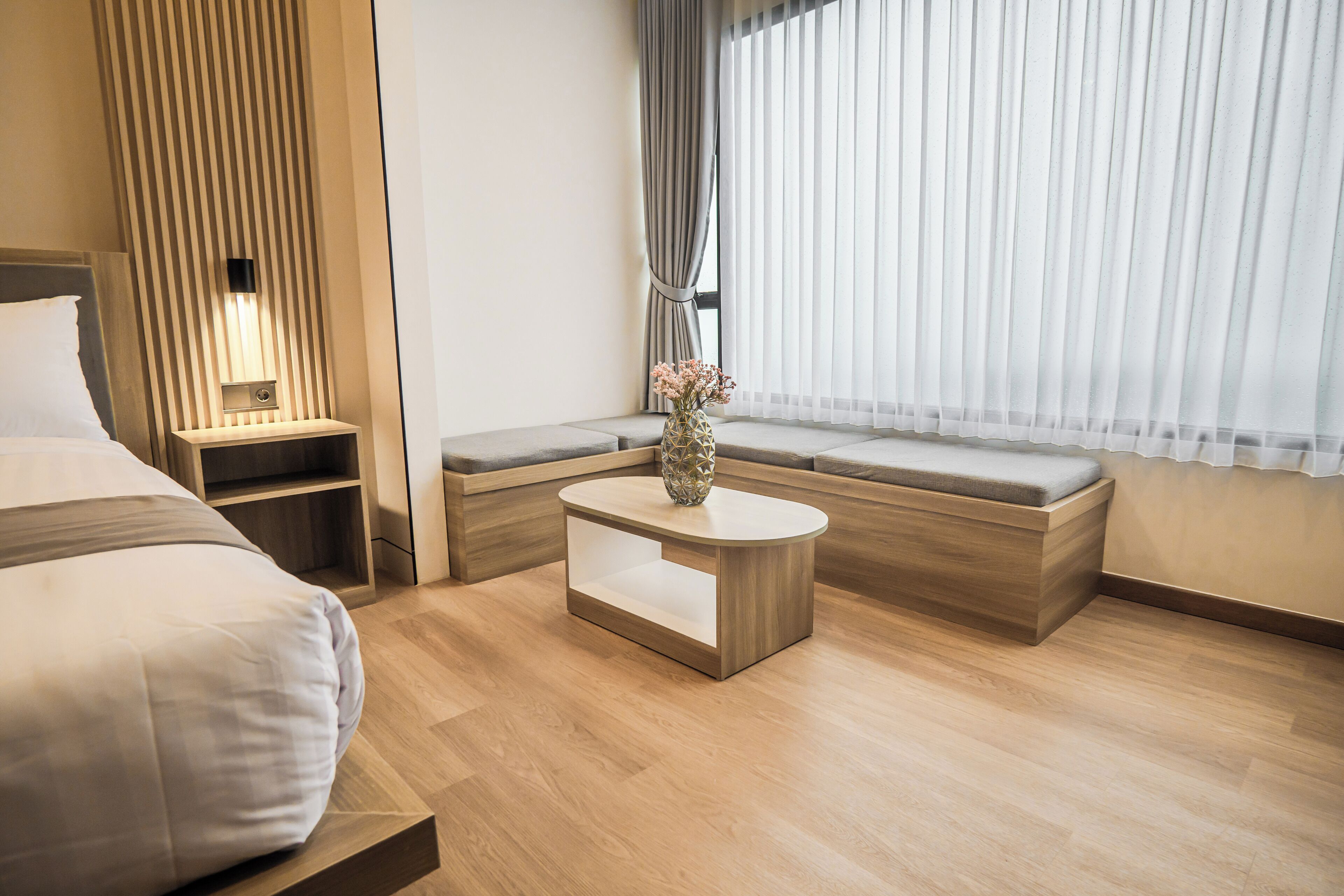A modern hotel room with a bed, bedside table, nightstand, and a seating area with a bench and a small table.