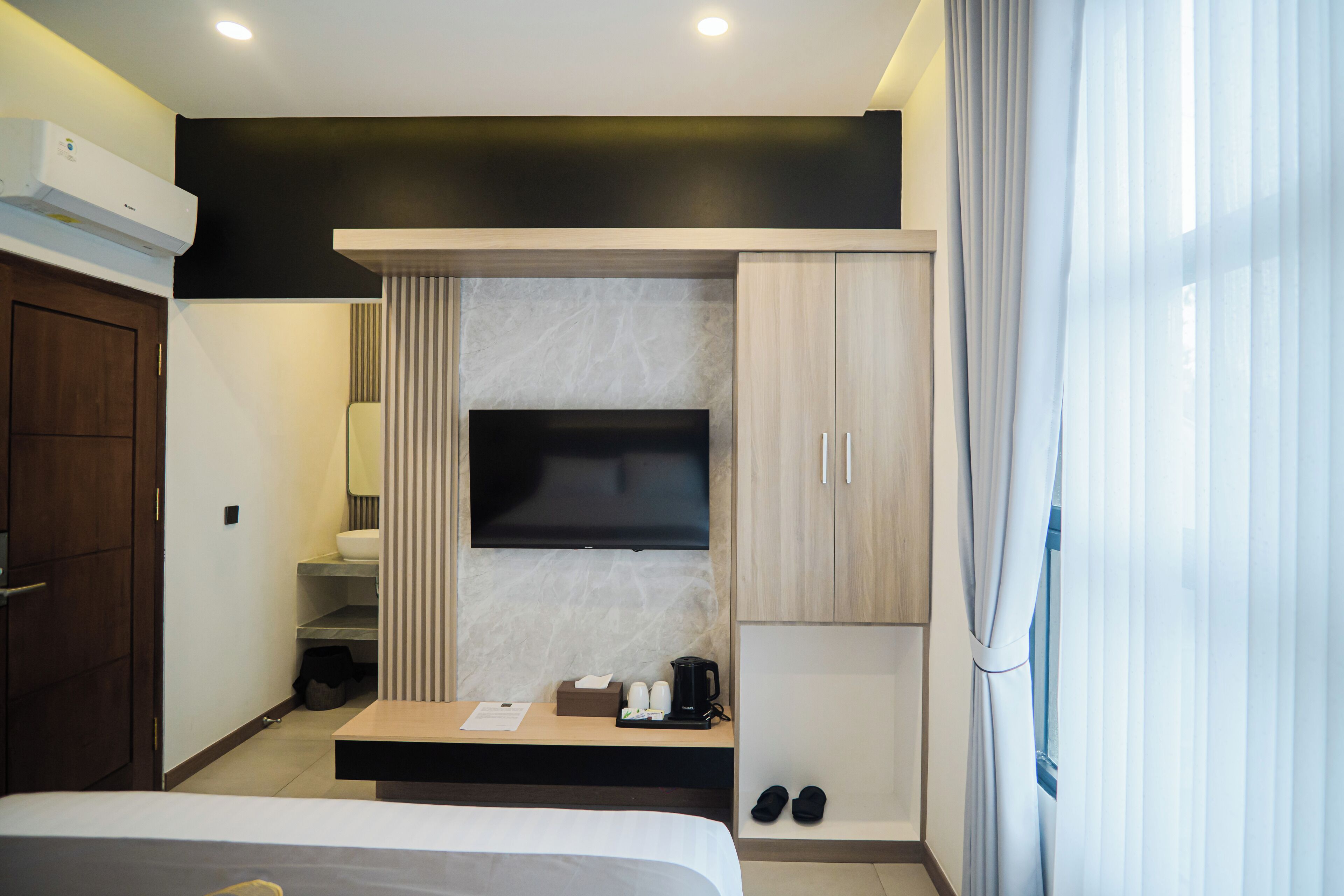 A modern hotel room with a bed, a flat-screen TV mounted on the wall, a coffee maker, slippers, and a small table with toiletries.