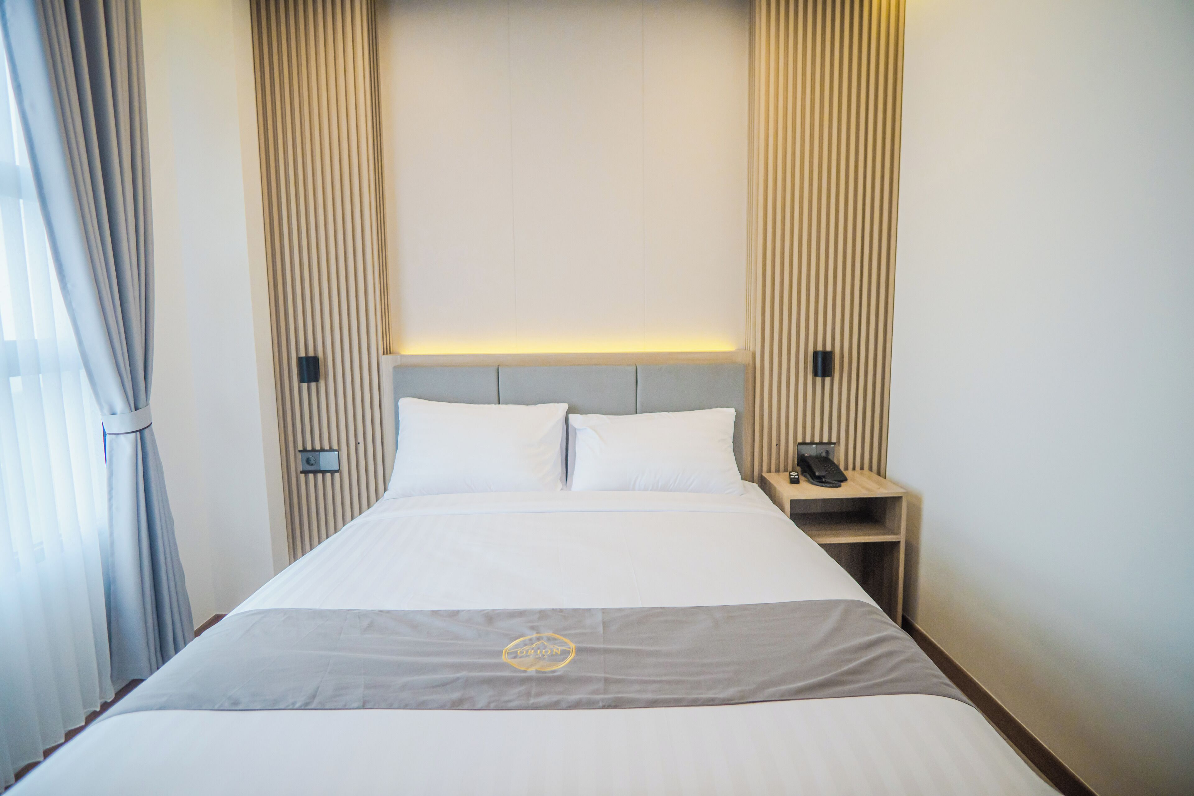 A neatly made bed with a grey and white bedspread, a headboard, and a bedside table with a lamp and phone.