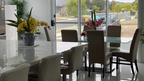 Un comedor moderno con una mesa y sillas de vidrio, un florero con flores amarillas, y vista a una calle residencial a través de una puerta de vidrio.