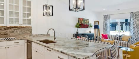 Une cuisine moderne avec un plan de travail en marbre, des meubles blancs et un îlot central doté de tabourets de bar. On y trouve également des luminaires suspendus et un tableau accroché au mur.