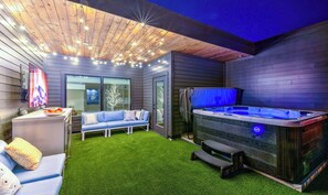 Un espace de vie extérieur moderne comprenant un jacuzzi, du gazon synthétique, un coin salon avec un canapé et des coussins, ainsi qu’un plafond en bois agrémenté de guirlandes lumineuses.